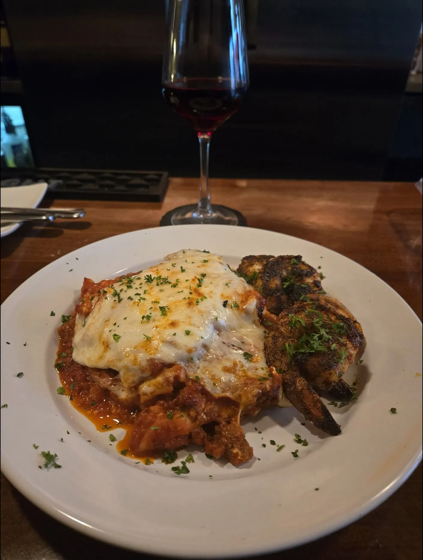 Lasagna beside blackened shrimp, with a glass of red wine behind.